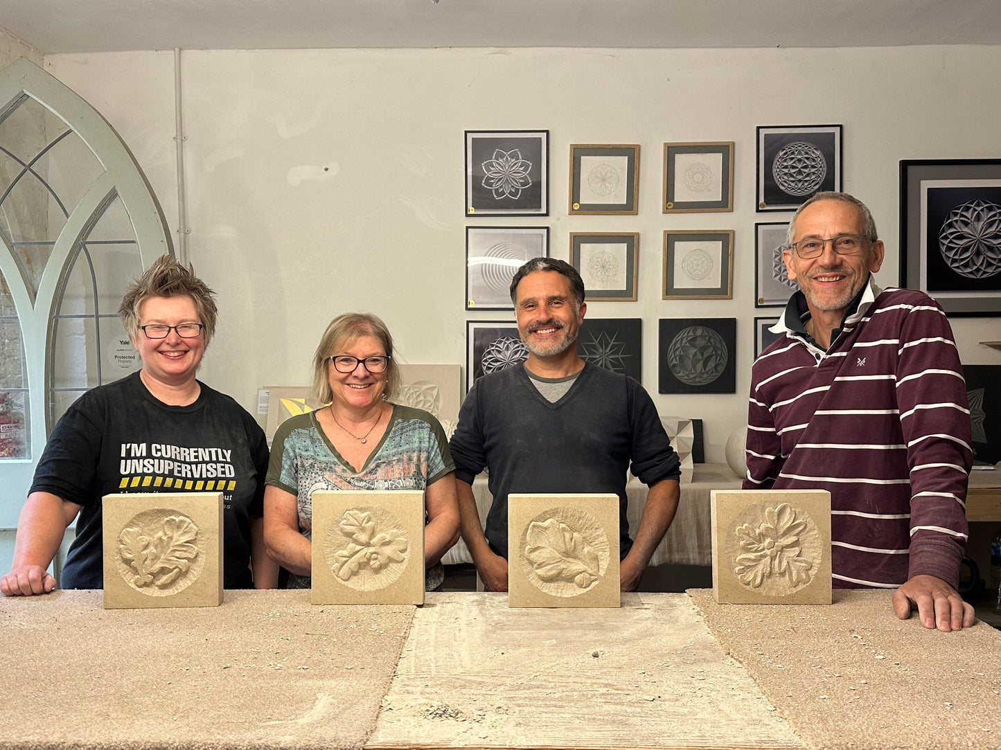 Four people smile while standing behind a table showcasing four beautifully carved stone panels with floral designs, presumably created during an inspiring course from Zoë Wilson Carving. The background is adorned with framed artworks, and one person sports a shirt with humorous text.