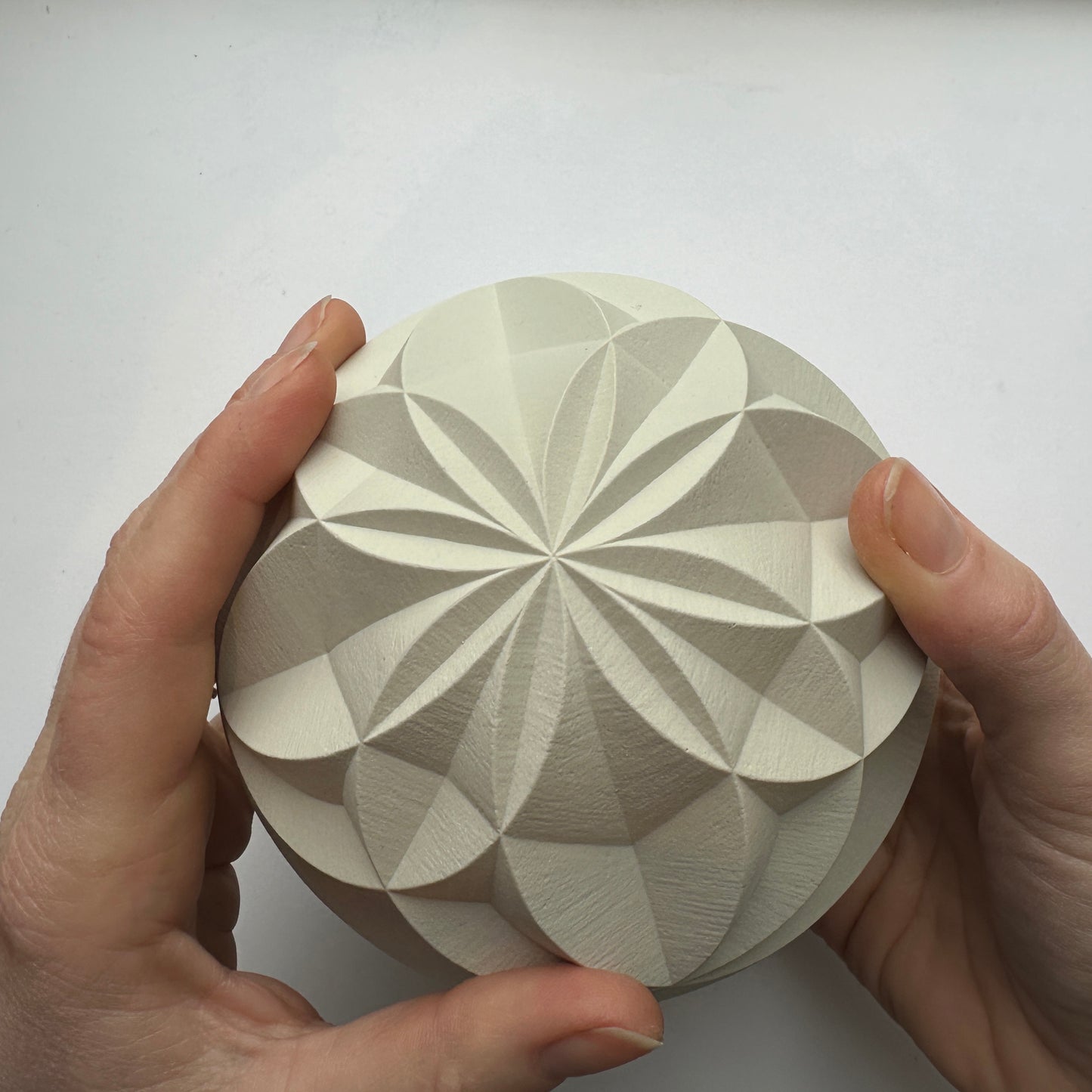 A hands-on view of The Canford Sphere, an intricately carved Jesmonite cast by Zoë Wilson Sculpture featuring geometric patterns that resemble petals or star shapes. This limited edition sculpture is crafted from a smooth, light-colored material.