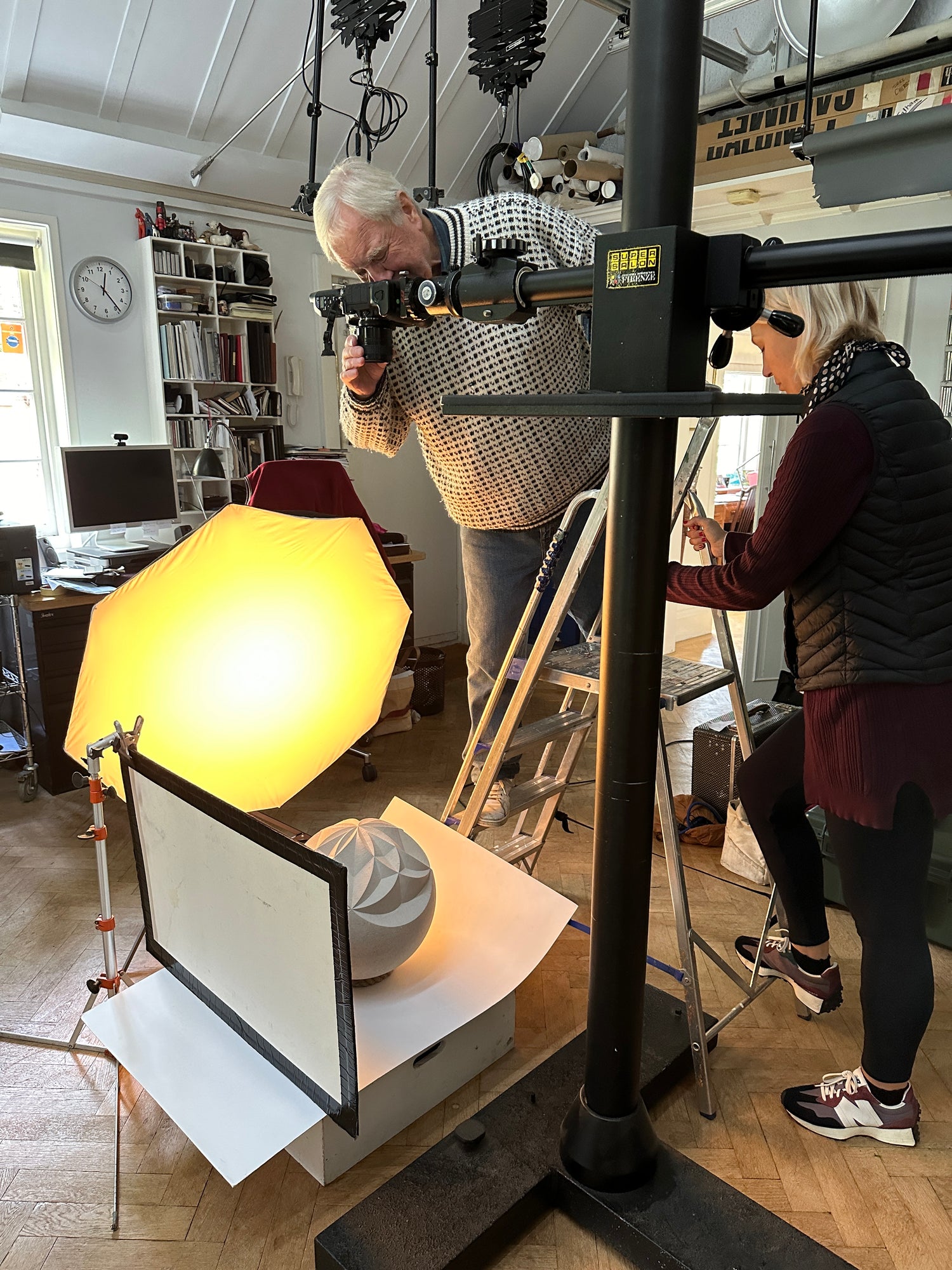 White haired man standing on a small step ladder looking down a camera lens in a photography studio. underneath the camera is one of Zoë's carved stone spheres