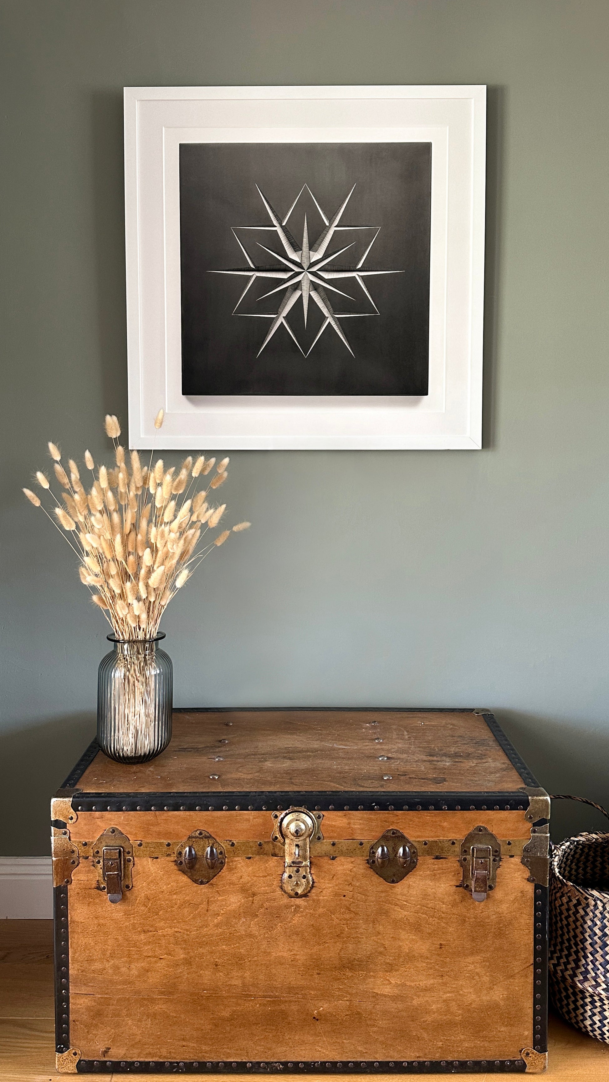 A rustic wooden chest with metal accents sits against a green wall. On top of the chest, a glass vase holds dried wheat stalks. Above it, "Snowflake," a fine art print by renowned photographer Julian Calder from Zoë Wilson Sculpture, features a star-like geometric design and hangs neatly.